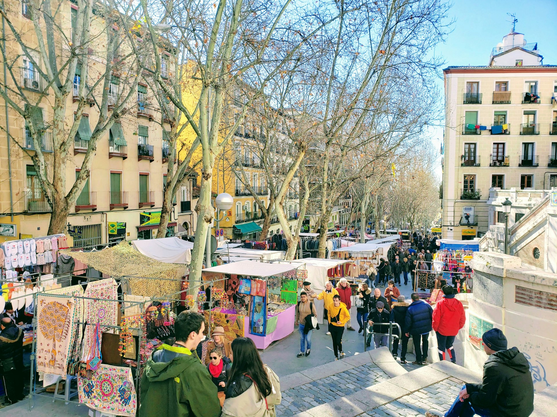 A vibrant street market bustling with locals and tourists, colorful stalls filled with handmade crafts and fresh fruits under a bright blue sky.