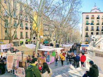 A vibrant outdoor market bustling with people. Colorful stalls with various items, including textiles and accessories, line the street. The market is surrounded by tall, leafless trees and residential buildings with multiple floors. People are seen walking, browsing, and interacting with each other under a clear blue sky.
