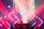 A DJ performs on stage surrounded by an array of geometric LED lights in vibrant red and blue tones. The lighting creates an energetic atmosphere, with beams and spotlights cutting through atmospheric haze. A silhouetted figure stands behind a mixing console, contributing to the vivid concert setting.