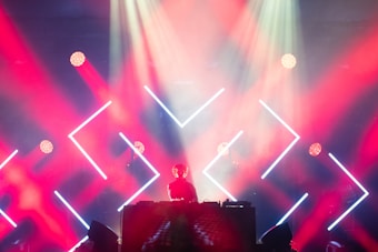 A DJ performs on stage surrounded by an array of geometric LED lights in vibrant red and blue tones. The lighting creates an energetic atmosphere, with beams and spotlights cutting through atmospheric haze. A silhouetted figure stands behind a mixing console, contributing to the vivid concert setting.