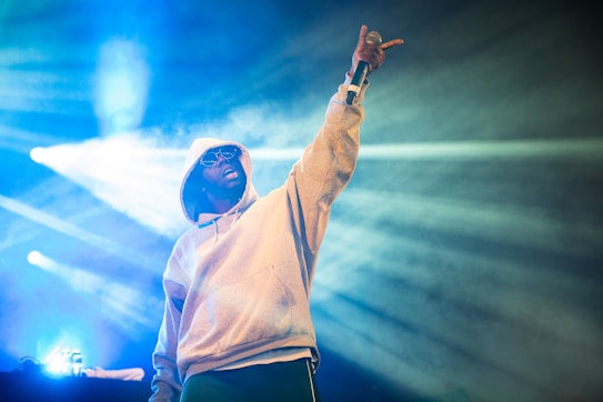 A person wearing a hooded sweatshirt and sunglasses is holding a microphone while pointing upward. The background features bright stage lights and smoke effects, creating a vibrant atmosphere.