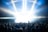 A large crowd of people is gathered in front of a brightly lit stage, with beams of light shining down from above. Many attendees have their hands raised, suggesting an energetic and lively event. The scene is set within an enclosed space, possibly under a tent or canopy.