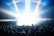 A large crowd of people is gathered in front of a brightly lit stage, with beams of light shining down from above. Many attendees have their hands raised, suggesting an energetic and lively event. The scene is set within an enclosed space, possibly under a tent or canopy.