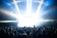 A large crowd of people is gathered in front of a brightly lit stage, with beams of light shining down from above. Many attendees have their hands raised, suggesting an energetic and lively event. The scene is set within an enclosed space, possibly under a tent or canopy.