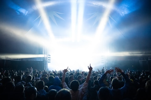 A large crowd of people is gathered in front of a brightly lit stage, with beams of light shining down from above. Many attendees have their hands raised, suggesting an energetic and lively event. The scene is set within an enclosed space, possibly under a tent or canopy.