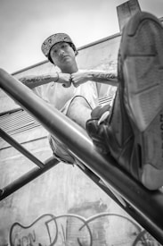 A young person is sitting on a metal structure, wearing a patterned cap and white shirt. The image is in black and white, giving it a dramatic tone. The perspective is from a low angle, emphasizing the youth's relaxed posture with legs extended and arms resting casually. Graffiti is visible on a wall in the background, contributing to an urban setting.
