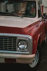 A close-up of the front section of a vintage red and white pickup truck, highlighting the headlight, grille, and part of the windshield. The vehicle shows signs of wear, with some rust visible on the bumper. The focus is primarily on the headlight and grille area.
