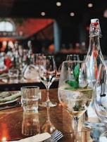 A restaurant table is set with clear wine glasses, a bottle of sparkling water, and a glass filled with a beverage garnished with mint leaves. The background is softly blurred, showcasing more wine glasses and dim lighting, creating a warm, inviting atmosphere.