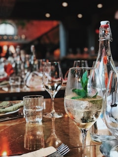 A restaurant table is set with clear wine glasses, a bottle of sparkling water, and a glass filled with a beverage garnished with mint leaves. The background is softly blurred, showcasing more wine glasses and dim lighting, creating a warm, inviting atmosphere.