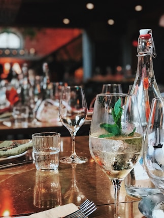 A restaurant table is set with clear wine glasses, a bottle of sparkling water, and a glass filled with a beverage garnished with mint leaves. The background is softly blurred, showcasing more wine glasses and dim lighting, creating a warm, inviting atmosphere.