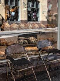 A bakery display window filled with a variety of rustic breads. Two vintage metal chairs are placed outside on the pavement, adding to the quaint atmosphere. The reflection of the street and buildings can be seen on the glass, enhancing the cozy feel of a small town bakery.