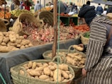 A market stall displaying a variety of fresh produce, including potatoes, onions, and tomatoes, arranged in woven baskets and crates. A person in traditional attire is selecting potatoes from a basket. In the background, more produce like oranges and grapes can be seen, with other people browsing the market.