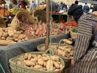 A market stall displaying a variety of fresh produce, including potatoes, onions, and tomatoes, arranged in woven baskets and crates. A person in traditional attire is selecting potatoes from a basket. In the background, more produce like oranges and grapes can be seen, with other people browsing the market.
