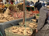 A market stall displaying a variety of fresh produce, including potatoes, onions, and tomatoes, arranged in woven baskets and crates. A person in traditional attire is selecting potatoes from a basket. In the background, more produce like oranges and grapes can be seen, with other people browsing the market.
