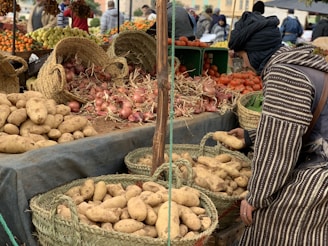 A market stall displaying a variety of fresh produce, including potatoes, onions, and tomatoes, arranged in woven baskets and crates. A person in traditional attire is selecting potatoes from a basket. In the background, more produce like oranges and grapes can be seen, with other people browsing the market.