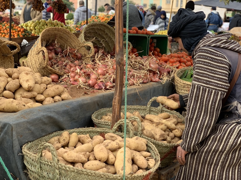 A market stall displaying a variety of fresh produce, including potatoes, onions, and tomatoes, arranged in woven baskets and crates. A person in traditional attire is selecting potatoes from a basket. In the background, more produce like oranges and grapes can be seen, with other people browsing the market.