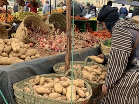 A market stall displaying a variety of fresh produce, including potatoes, onions, and tomatoes, arranged in woven baskets and crates. A person in traditional attire is selecting potatoes from a basket. In the background, more produce like oranges and grapes can be seen, with other people browsing the market.