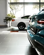 Two luxury cars are parked inside a well-lit showroom with large windows and a view of trees outside. The cars appear to have a sleek, modern design, and the setting includes tiled flooring and plants.