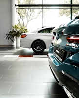 Two luxury cars are parked inside a well-lit showroom with large windows and a view of trees outside. The cars appear to have a sleek, modern design, and the setting includes tiled flooring and plants.