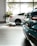 Two luxury cars are parked inside a well-lit showroom with large windows and a view of trees outside. The cars appear to have a sleek, modern design, and the setting includes tiled flooring and plants.