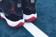 A pair of black and white sneakers with red soles placed on a blue outdoor sports court. The shoes feature a distinct red silhouette logo on the back. The court shows a white line separating two sections.