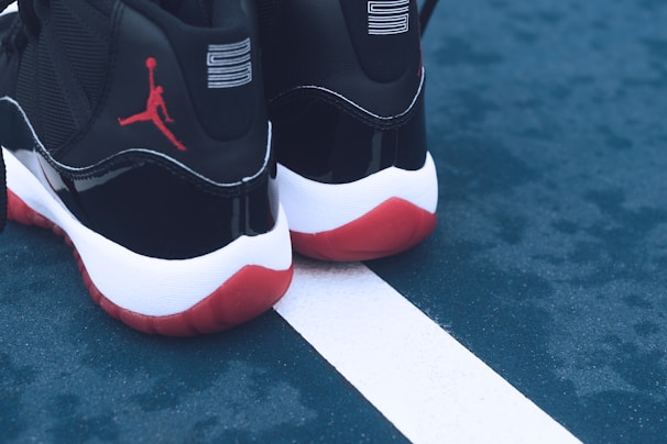 A pair of black and white sneakers with red soles placed on a blue outdoor sports court. The shoes feature a distinct red silhouette logo on the back. The court shows a white line separating two sections.