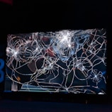 A large, cracked and shattered glass surface with intricate patterns of fractures spreading out in a web-like structure. The glass is reflective, catching some light, creating bright spots and highlights amidst the dark background.