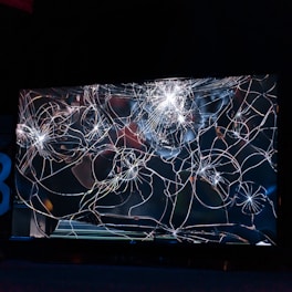 A large, cracked and shattered glass surface with intricate patterns of fractures spreading out in a web-like structure. The glass is reflective, catching some light, creating bright spots and highlights amidst the dark background.