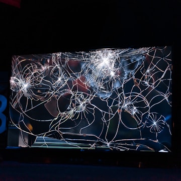 A large, cracked and shattered glass surface with intricate patterns of fractures spreading out in a web-like structure. The glass is reflective, catching some light, creating bright spots and highlights amidst the dark background.