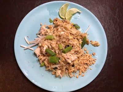 A plate of pad thai garnished with green onion, crushed peanuts, and lime wedges is on a blue plate. It includes rice noodles, pieces of chicken or tofu, and is sprinkled with red chili flakes.