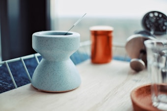 A modern kitchen countertop with a blue, speckled ceramic mug featuring a unique hourglass shape. A metal stirrer rests inside the mug. In the background, there is an orange tin canister, a French press coffee maker with a glass and metal design, and a clear glass cup on an orange coaster, all placed on a wooden surface with a metal railing visible.