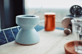 A modern kitchen countertop with a blue, speckled ceramic mug featuring a unique hourglass shape. A metal stirrer rests inside the mug. In the background, there is an orange tin canister, a French press coffee maker with a glass and metal design, and a clear glass cup on an orange coaster, all placed on a wooden surface with a metal railing visible.
