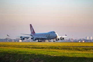 An airplane labeled 'Cargolux Italia' is taxiing on a runway at an airport. The aircraft is a large cargo plane with four engines, displaying distinctive red and white coloring on its tail. The scene is set against a clear sky with a vast expanse of grassy field in the foreground and distant buildings and wind turbines in the background.