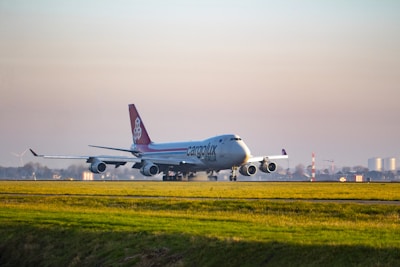 An airplane labeled 'Cargolux Italia' is taxiing on a runway at an airport. The aircraft is a large cargo plane with four engines, displaying distinctive red and white coloring on its tail. The scene is set against a clear sky with a vast expanse of grassy field in the foreground and distant buildings and wind turbines in the background.