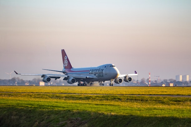 An airplane labeled 'Cargolux Italia' is taxiing on a runway at an airport. The aircraft is a large cargo plane with four engines, displaying distinctive red and white coloring on its tail. The scene is set against a clear sky with a vast expanse of grassy field in the foreground and distant buildings and wind turbines in the background.