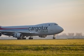A large cargo airplane is seen taxiing on a runway surrounded by green grass. The plane is branded with the logo 'Cargolux Italia' on its body. The sky is overcast with soft sunlight creating a calm ambiance. In the background, there are silhouettes of trees and possibly a distant building.