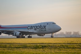 A large cargo airplane is seen taxiing on a runway surrounded by green grass. The plane is branded with the logo 'Cargolux Italia' on its body. The sky is overcast with soft sunlight creating a calm ambiance. In the background, there are silhouettes of trees and possibly a distant building.