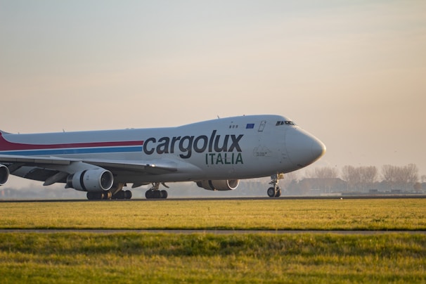 A large cargo airplane is seen taxiing on a runway surrounded by green grass. The plane is branded with the logo 'Cargolux Italia' on its body. The sky is overcast with soft sunlight creating a calm ambiance. In the background, there are silhouettes of trees and possibly a distant building.