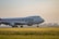 A large cargo airplane is seen taxiing on a runway surrounded by green grass. The plane is branded with the logo 'Cargolux Italia' on its body. The sky is overcast with soft sunlight creating a calm ambiance. In the background, there are silhouettes of trees and possibly a distant building.