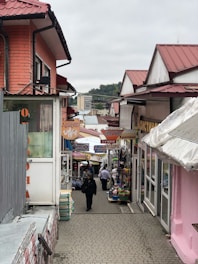 A narrow, bustling market street flanked by colorful shops with red roofs. Signs with Cyrillic script hang above the path, and multiple people walk along the cobblestone path lined with goods and stalls.