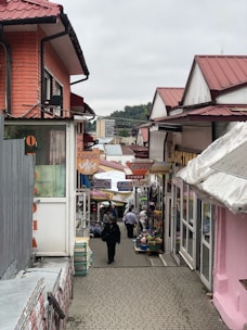 A narrow, bustling market street flanked by colorful shops with red roofs. Signs with Cyrillic script hang above the path, and multiple people walk along the cobblestone path lined with goods and stalls.