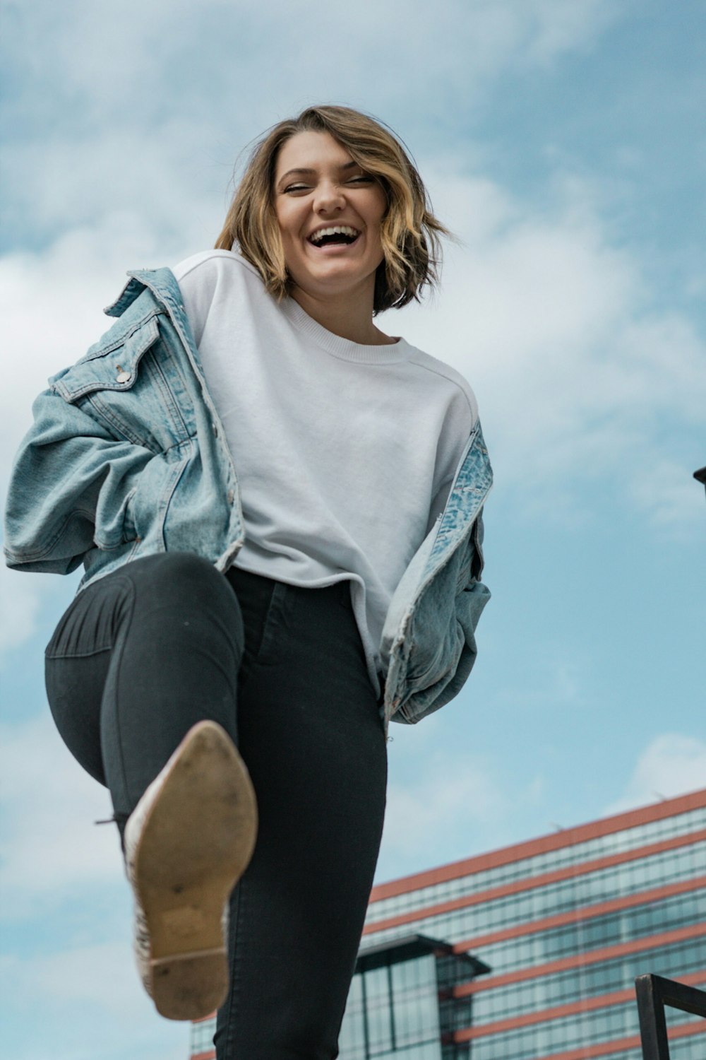 woman wearing white crew-neck t-shirt and blue denim jacket laughing while lifting right leg