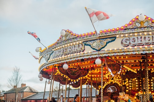 A vibrant carousel with Victorian-style decorations and intricate lighting stands in an outdoor setting. The top is adorned with colorful patterns and a striped flag fluttering in the breeze. Nearby, brick buildings and leafless trees can be seen under a cloudy sky.
