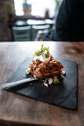 A beautifully plated dish featuring a slice of toasted bread topped with shredded meat, garnished with a poached egg and fresh cilantro. The meal is served on a black slate plate with sliced radishes and green herbs scattered around. Natural light illuminates the rustic wooden table, creating a cozy dining atmosphere.