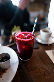 A glass filled with a vibrant red beverage is placed on a wooden table. The drink is topped with ice and accompanied by a blue and white striped straw. Nearby, there is a small metallic cup on a white plate, holding some sauce. In the blurred background, a cup of coffee is visible alongside some greenery, possibly a salad.