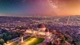 A prominent observatory stands on a hill overlooking a sprawling urban landscape during sunset. The sky is filled with stars and a faint glow of the Milky Way, creating a stunning celestial backdrop. The city lights begin to illuminate the densely packed buildings below, while the observatory is surrounded by well-manicured greenery and pathways.