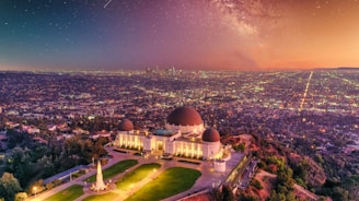 A prominent observatory stands on a hill overlooking a sprawling urban landscape during sunset. The sky is filled with stars and a faint glow of the Milky Way, creating a stunning celestial backdrop. The city lights begin to illuminate the densely packed buildings below, while the observatory is surrounded by well-manicured greenery and pathways.