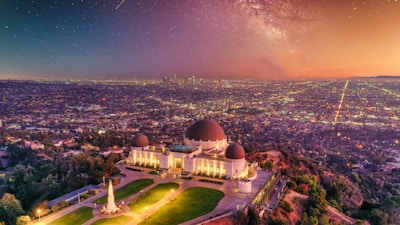 A prominent observatory stands on a hill overlooking a sprawling urban landscape during sunset. The sky is filled with stars and a faint glow of the Milky Way, creating a stunning celestial backdrop. The city lights begin to illuminate the densely packed buildings below, while the observatory is surrounded by well-manicured greenery and pathways.