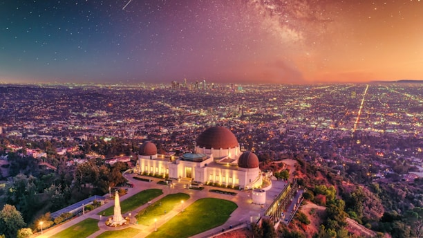 A prominent observatory stands on a hill overlooking a sprawling urban landscape during sunset. The sky is filled with stars and a faint glow of the Milky Way, creating a stunning celestial backdrop. The city lights begin to illuminate the densely packed buildings below, while the observatory is surrounded by well-manicured greenery and pathways.
