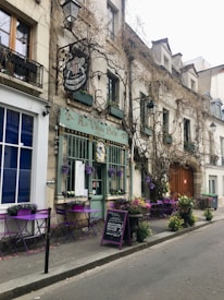 A charming Parisian street café with vintage appeal features purple tables and chairs aligned outside. Climbing vines decorate the building's exterior, and colorful flowers add a welcoming touch.